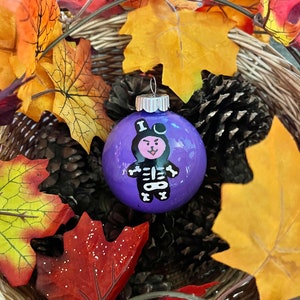 May include: Purple glass Christmas ornament with a black and white skeleton wearing a witch's hat. The ornament is surrounded by fall leaves.