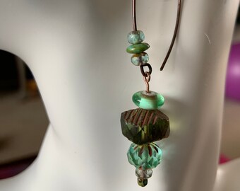 May include: A pair of green and brown dangle earrings with a copper wire hoop. The earrings feature a variety of green glass beads, including a faceted bead, a round bead, and a rectangular bead.