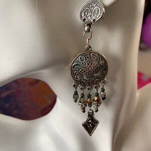 May include: A silver-toned earring with an ornate design. The earring features a heart-shaped top, a circular center with intricate patterns, and dangling beads in shades of green and gold. A diamond-shaped charm hangs at the bottom.
