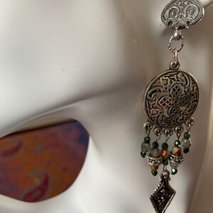 May include: A silver-toned earring with an ornate design. The earring features a heart-shaped top, a circular pendant with intricate patterns, and dangling beads in shades of green and iridescent colors. A diamond-shaped charm hangs at the bottom.