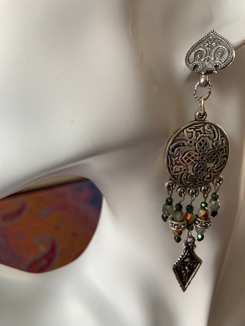 May include: A silver-toned earring with an ornate design. The earring features a heart-shaped top, a circular pendant with intricate patterns, and dangling beads in shades of green and iridescent colors. A diamond-shaped charm hangs at the bottom.