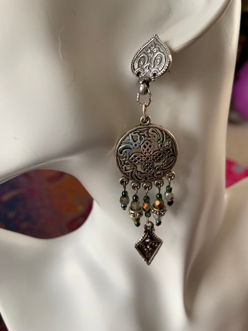 May include: A silver-toned earring with an ornate design. The earring features a heart-shaped top, a circular center with intricate patterns, and dangling beads in shades of green and gold. A diamond-shaped charm hangs at the bottom.