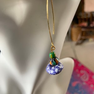 Op de afbeelding: Gouden oorhangers met blauwe en witte gepatroneerde keramische kralen en groene en oranje glazen kralen.