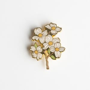 May include: A gold-toned metal pin featuring a cluster of white dogwood flowers with yellow centers and green leaves. The pin has a detailed design and is set against a white background, showcasing the floral arrangement.