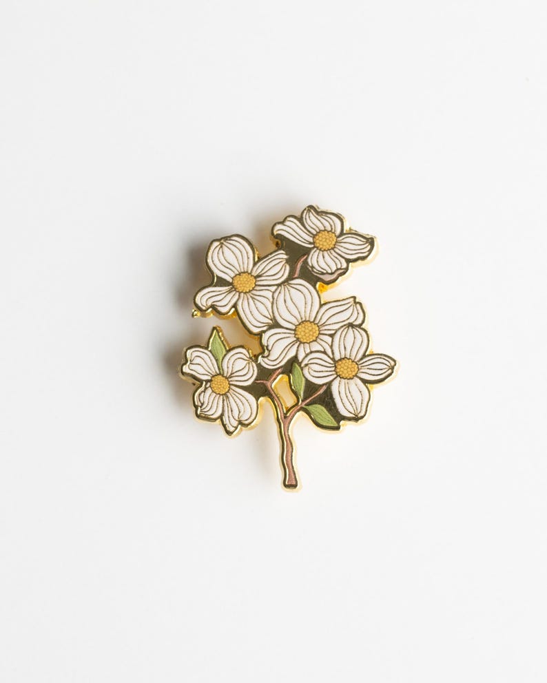May include: A gold-toned metal pin featuring a cluster of white dogwood flowers with yellow centers and green leaves. The pin has a detailed design and is set against a white background, showcasing the floral arrangement.
