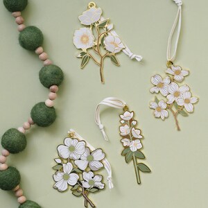 May include: Four floral-shaped ornaments with white flowers and gold-colored metal trim. Each ornament has a white ribbon for hanging. A green felt ball garland is in the background. The ornaments are on a light green surface.