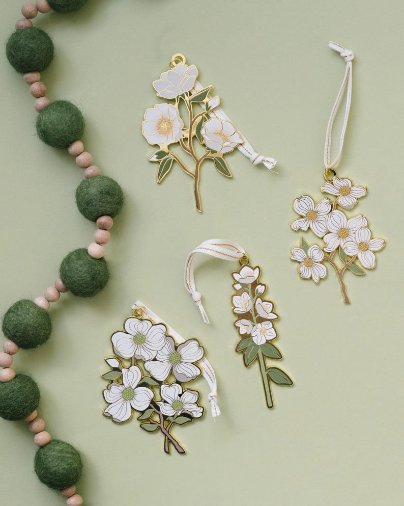 May include: Four floral-shaped ornaments with white flowers and gold-colored metal trim. Each ornament has a white ribbon for hanging. A green felt ball garland is in the background. The ornaments are on a light green surface.