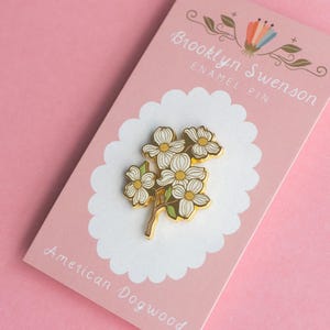 May include: An enamel pin featuring a cluster of white dogwood flowers with yellow centers and green leaves. The pin is gold-colored and set on a pink backing card with the text "Brooklyn Swenson ENAMEL PIN" and "American Dogwood".