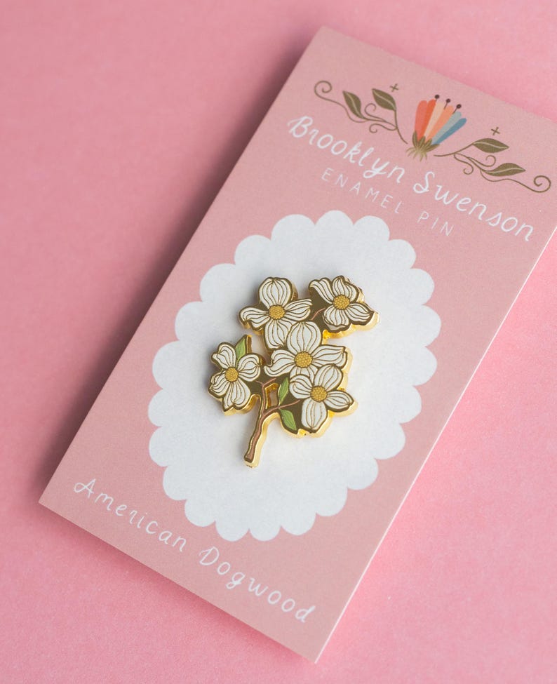 May include: An enamel pin featuring a cluster of white dogwood flowers with yellow centers and green leaves. The pin is gold-colored and set on a pink backing card with the text "Brooklyn Swenson ENAMEL PIN" and "American Dogwood".