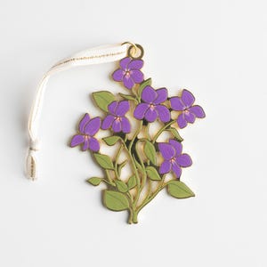 May include: A floral ornament featuring a cluster of purple violets with green leaves and gold-colored stems. The ornament has a cream-colored ribbon for hanging. The violets have a detailed design, and the overall aesthetic is delicate and nature-inspired.