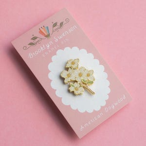 May include: An enamel pin featuring a cluster of white and yellow dogwood flowers with green accents. The pin is on a pink card with the text "Brooklyn Swenson ENAMEL PIN" and "American Dogwood".