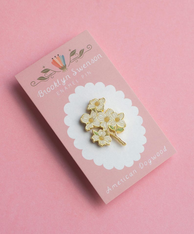 May include: An enamel pin featuring a cluster of white and yellow dogwood flowers with green accents. The pin is on a pink card with the text "Brooklyn Swenson ENAMEL PIN" and "American Dogwood".