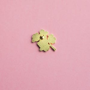 May include: A gold-colored metal pin shaped like a four-leaf clover. The pin has a textured surface and a small, round clasp on the back. The pin is set against a solid pink background.
