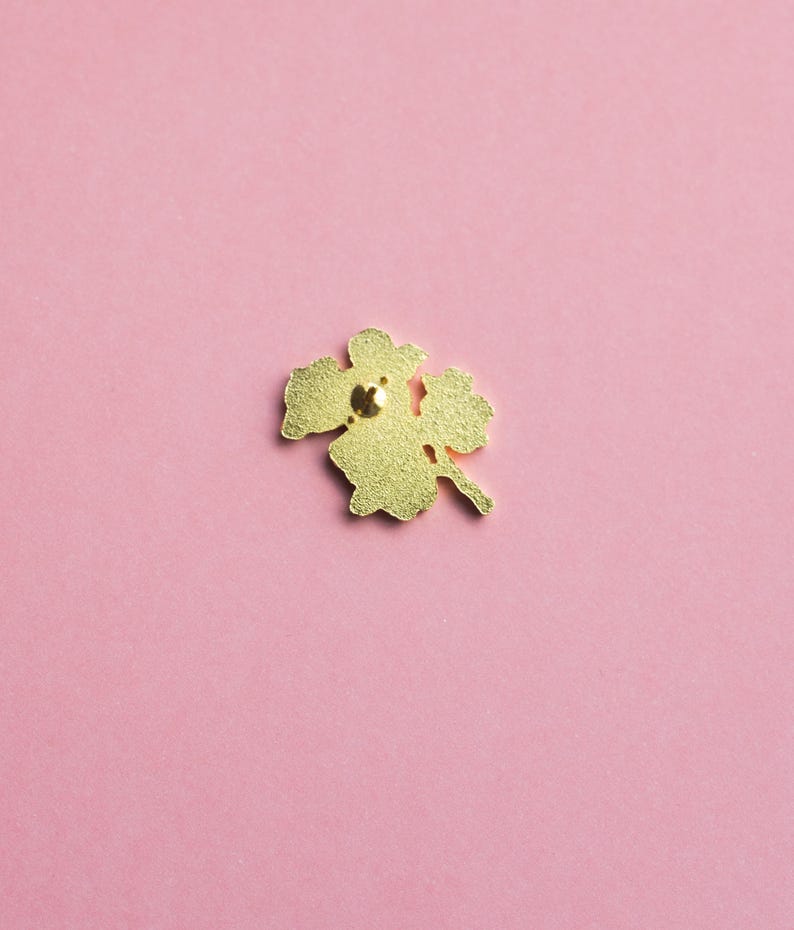 May include: A gold-colored metal pin shaped like a four-leaf clover. The pin has a textured surface and a small, round clasp on the back. The pin is set against a solid pink background.