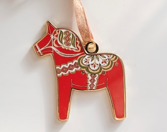 May include: A red Dala horse ornament with gold trim and a decorative floral design in white, green, and gold. The ornament has a gold ribbon for hanging. A festive decoration for the holidays.