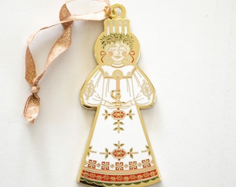 May include: Gold-colored metal ornament depicting a woman in a white dress with a red and gold patterned skirt, holding a candle. She is wearing a crown of candles and a green wreath.
