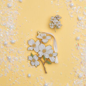 May include: Three floral-themed accessories on a yellow background. The accessories include a pin and a charm with white flowers, gold accents, and green stems. The charm has a cream-colored ribbon. The background is decorated with white confetti and pearls.