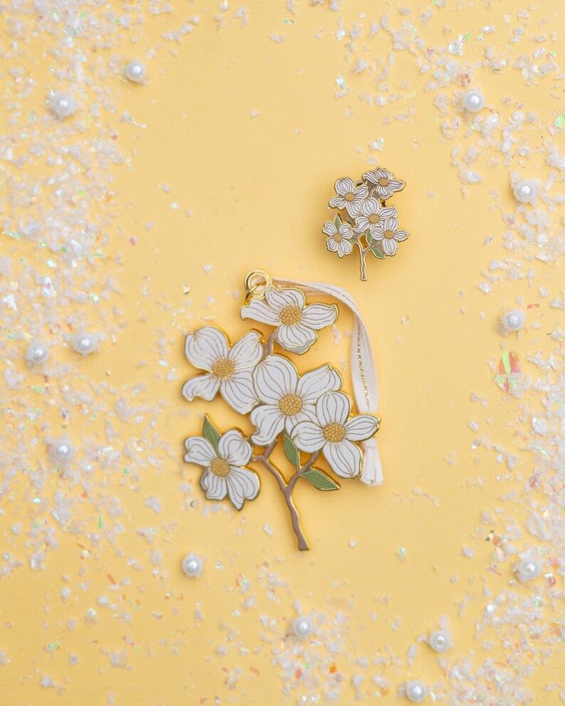May include: Three floral-themed accessories on a yellow background. The accessories include a pin and a charm with white flowers, gold accents, and green stems. The charm has a cream-colored ribbon. The background is decorated with white confetti and pearls.
