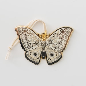 Puede incluir: Adorno decorativo con forma de mariposa con borde dorado y fondo crema. La mariposa presenta intrincados patrones florales y geométricos en negro, crema y dorado. Se adjunta una cinta color crema para colgar.