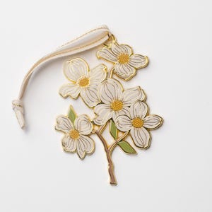 May include: A floral ornament featuring white flowers with gold outlines and yellow centers, attached to a beige ribbon. The ornament has green leaves and a brown stem, set against a white background. This decorative piece is suitable for seasonal decor.