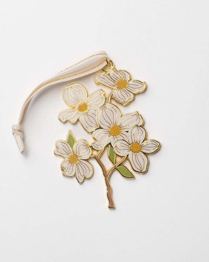 May include: A floral ornament featuring white flowers with gold outlines and yellow centers, attached to a beige ribbon. The ornament has green leaves and a brown stem, set against a white background. This decorative piece is suitable for seasonal decor.