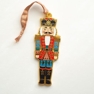 May include: A gold-colored metal Christmas ornament shaped like a nutcracker soldier. The ornament features a red jacket, blue pants, and a black hat with gold accents. A brown ribbon is attached for hanging.