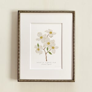 Puede incluir: Una impresi&oacute;n enmarcada de una flor de cornejo blanco con hojas verdes. La impresi&oacute;n tiene el texto "NORTH CAROLINA" y "American Dogwood" debajo de la imagen.