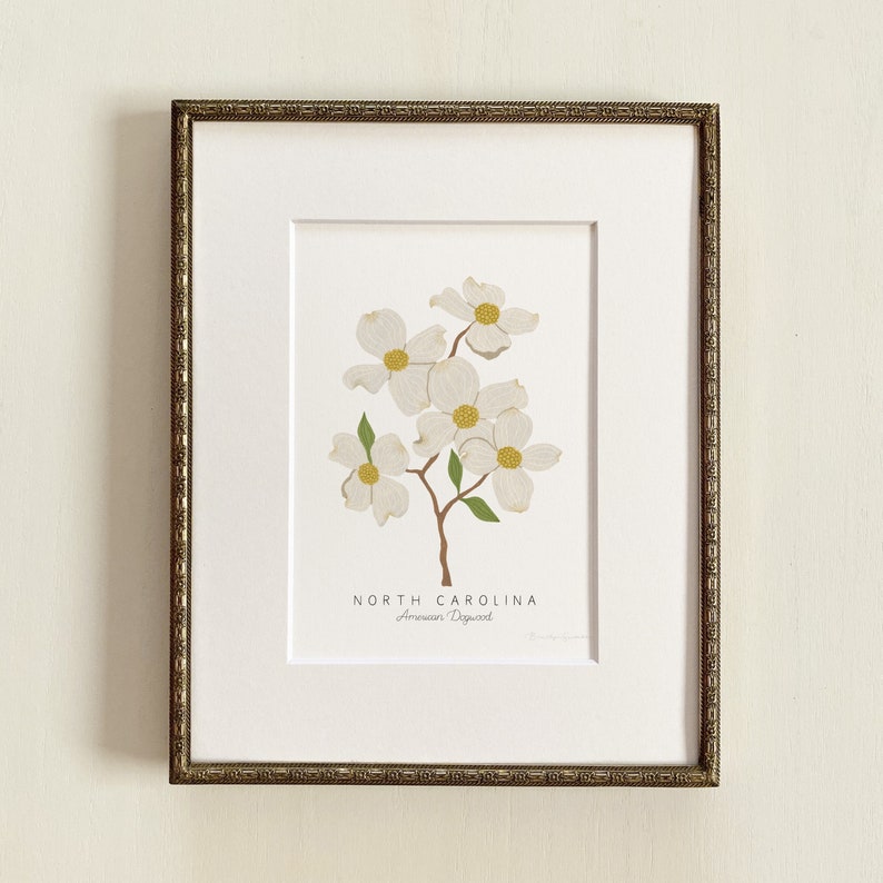 Puede incluir: Una impresi&oacute;n enmarcada de una flor de cornejo blanco con hojas verdes. La impresi&oacute;n tiene el texto "NORTH CAROLINA" y "American Dogwood" debajo de la imagen.
