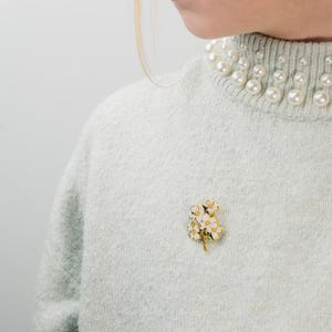 May include: A light blue sweater with a pearl-embellished collar. A gold-toned brooch with white and yellow flowers is pinned on the sweater. The brooch adds a touch of elegance to the outfit.