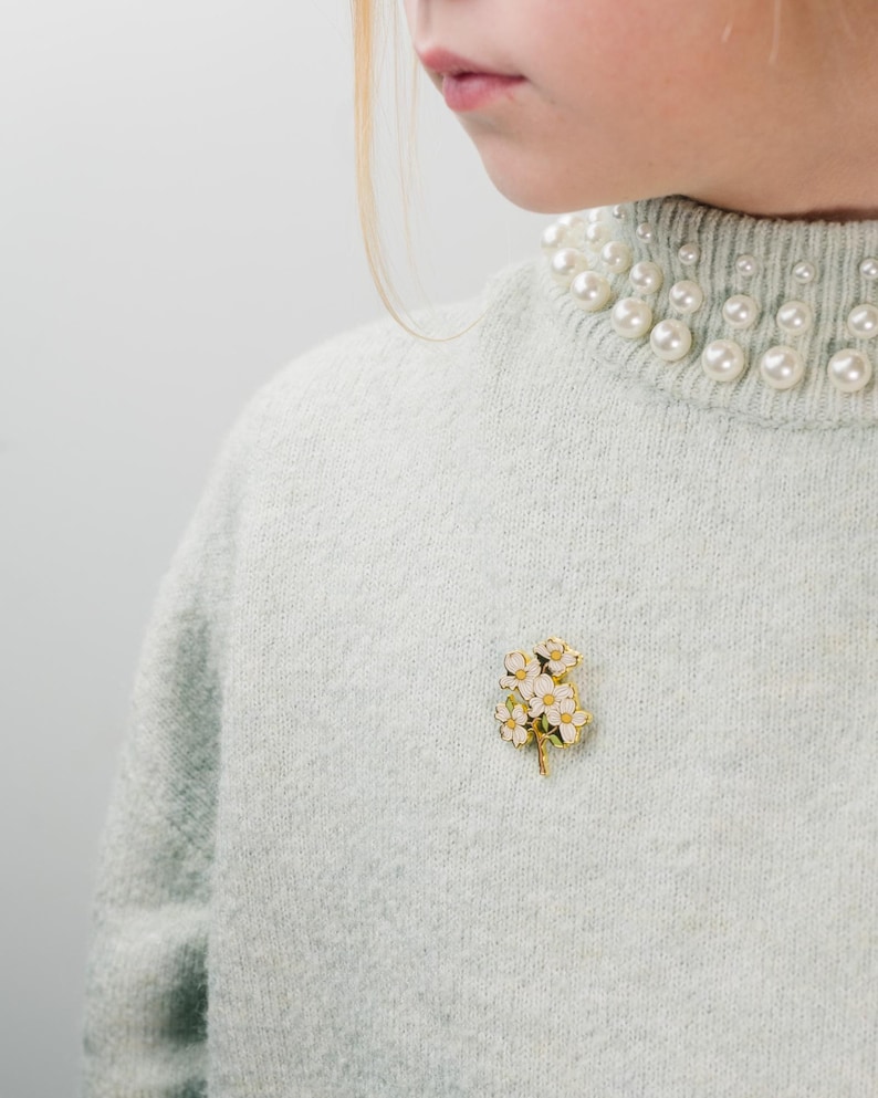 May include: A light blue sweater with a pearl-embellished collar. A gold-toned brooch with white and yellow flowers is pinned on the sweater. The brooch adds a touch of elegance to the outfit.