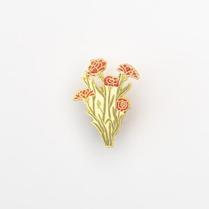 May include: A gold-colored enamel pin featuring a bouquet of red carnations with green stems and leaves. The pin has a detailed design with a gold outline, and the flowers are in full bloom. The pin is set against a white background.