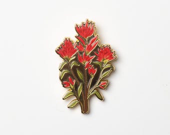 May include: A gold-toned metal pin featuring a cluster of red flowers with green leaves. The flowers have a detailed, textured appearance, and the pin has a polished finish. The design is set against a white background.