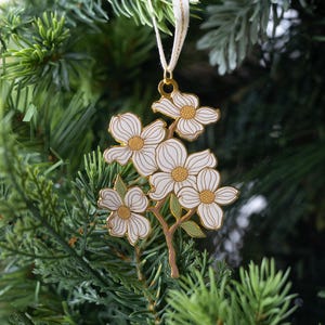 May include: A white and gold floral ornament with a cream-colored ribbon, hanging on a Christmas tree. The ornament features a cluster of white flowers with gold centers and outlines, and green leaves.