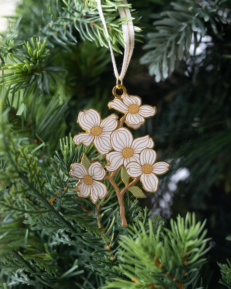 May include: A white and gold floral ornament with a cream-colored ribbon, hanging on a Christmas tree. The ornament features a cluster of white flowers with gold centers and outlines, and green leaves.
