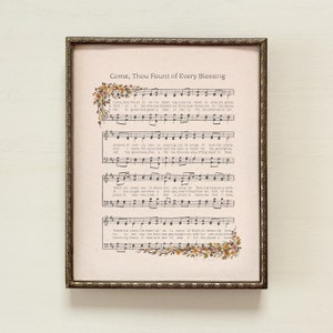 May include: Framed print featuring sheet music with the title "Come, Thou Fount of Every Blessing." The text is accompanied by musical notes and floral accents in the corners. The frame is a dark, ornate gold color.