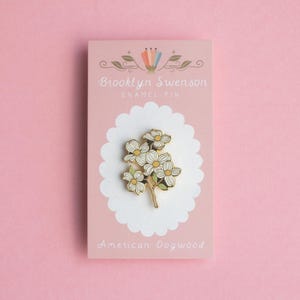 May include: An enamel pin featuring a cluster of white and yellow flowers with green stems, set against a pink card. The card reads "Brooklyn Swenson ENAMEL PIN" and "American Dogwood". The pin is on a white scalloped background.