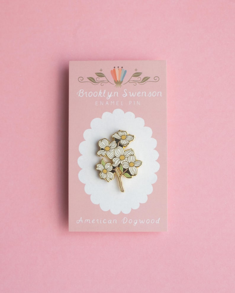 May include: An enamel pin featuring a cluster of white and yellow flowers with green stems, set against a pink card. The card reads "Brooklyn Swenson ENAMEL PIN" and "American Dogwood". The pin is on a white scalloped background.