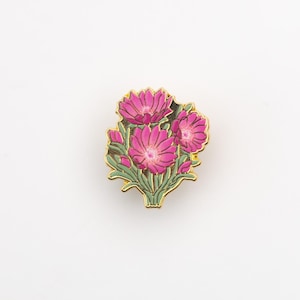 Puede incluir: Un broche de metal dorado con un racimo de flores rosas con hojas verdes. Las flores tienen pétalos detallados y están dispuestas en forma de ramo. El broche tiene un borde decorativo detallado.