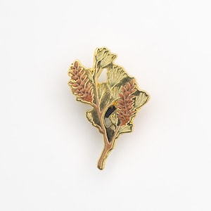 May include: A gold-toned metal pin featuring a botanical design. The pin depicts a stylized branch with leaves and seed pods in shades of gold, brown, and green. The pin is set against a white background.