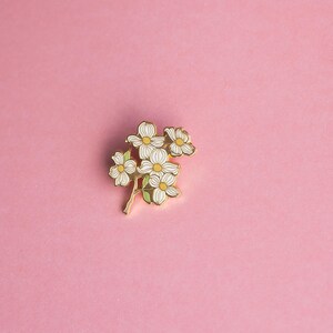 May include: A gold-colored floral enamel pin featuring white flowers with yellow centers and green leaves. The pin is set against a solid pink background, creating a striking contrast. This decorative pin is perfect for adding a touch of nature-inspired flair to any outfit.