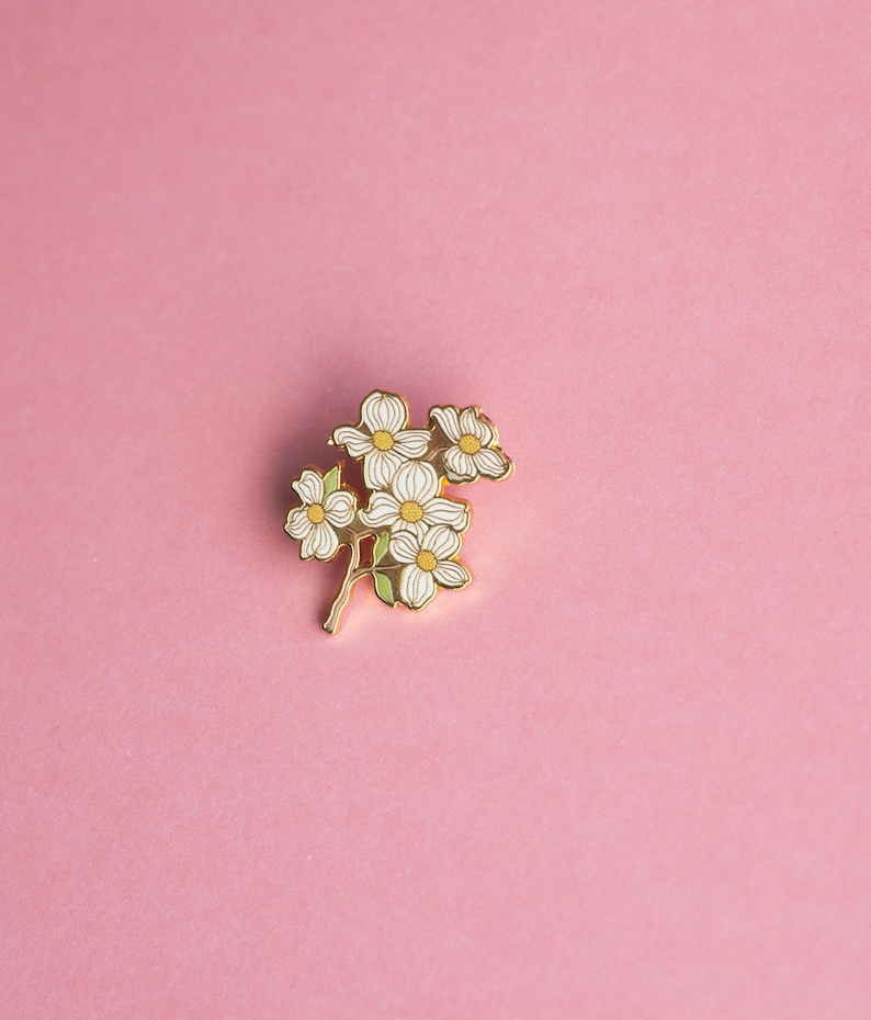 May include: A gold-colored floral enamel pin featuring white flowers with yellow centers and green leaves. The pin is set against a solid pink background, creating a striking contrast. This decorative pin is perfect for adding a touch of nature-inspired flair to any outfit.