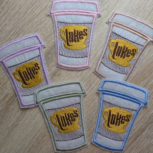 May include: Five embroidered patches in the shape of coffee cups. Each patch features the word "Lukes" in a yellow, stylized font, above a yellow coffee cup design. The patches have different colored borders: pink, purple, beige, green, and blue.