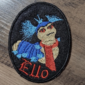 May include: Embroidered patch with a cartoon character with blue hair, red eyes, and a red scarf. The character is wearing a blue and white outfit and the patch says "Ello" in red lettering.