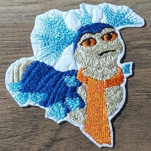 May include: Embroidered patch featuring a whimsical creature with blue, orange, and white details. The design includes a blue head, orange scarf, and a blue and white patterned body. The patch is on a wooden surface.