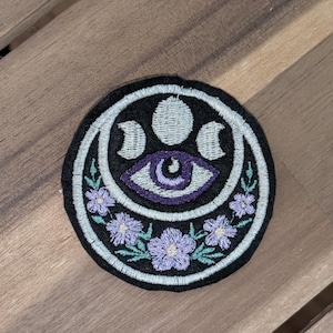 May include: Embroidered patch featuring a celestial design. The patch has a black background with a light blue border. The design includes a purple eye, crescent moons, and light purple flowers. The patch is round.