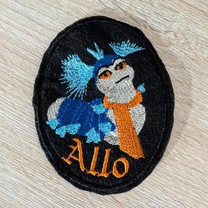 May include: Embroidered patch featuring a whimsical creature with blue and silver details, an orange scarf, and the word "Allo" in orange. The patch has a black oval border.
