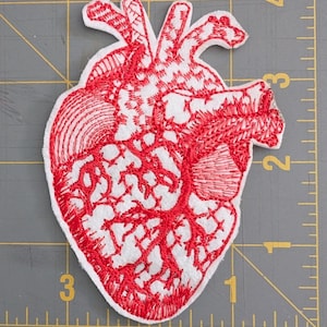 May include: Embroidered patch in the shape of a human heart. The heart is white with red embroidery detailing the veins and outer shape. The patch is on a gray surface with a ruler.