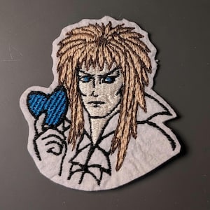 May include: Embroidered patch featuring a stylized portrait of a character with long, light brown hair and blue eyes. The character is holding a blue heart. The patch has a white border and is on a gray background.