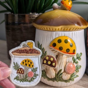 May include: A white ceramic mushroom-shaped cookie jar with a brown lid and a yellow mushroom design. A small embroidered patch with a mushroom design is in the foreground.