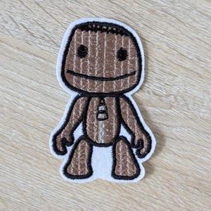 May include: A brown and tan embroidered patch featuring a cartoon character with a stitched appearance. The character has a smiling face, black eyes, and a white border. The patch is designed to be ironed on or sewn onto fabric.